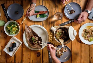 People with chopsticks enjoying various Japanese dishes placed on a wooden table.