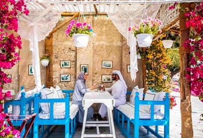 A couple in traditional Emirati attire dining at the vibrant, flower-filled outdoor courtyard of Arabian Tea House