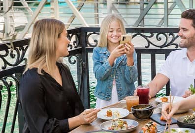 A woman sitting in front of her sushi and smiling while looking at a little girl by her side that is taking picture of the table and also smiling