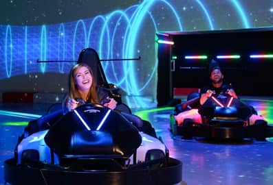 A woman happily driving and chaos kart in a closed space with laser projections in the background