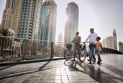 Two cyclers with their bikes, strolling around Dubai Marina Walk in day time, with residential builds in the background