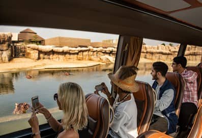 Visitors on a bus passing by a water channel with hippos while taking photographs.