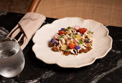 A plated gourmet dish at Sufret Maryam topped with fresh vegetables and sauce, served on a white floral-shaped plate over a dark marble surface