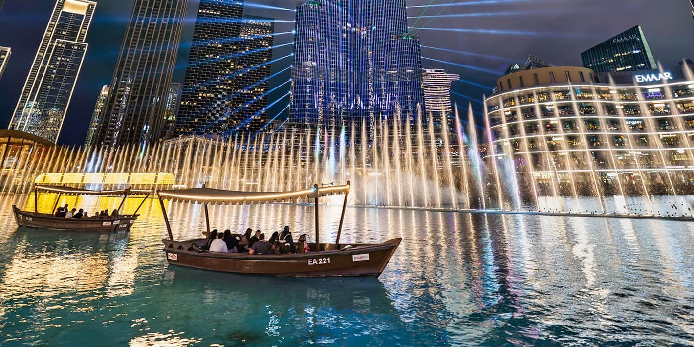 Panoramic view of the Dubai Fountain at night with Abras in the foreground and Burj Khalifa and its laser show in the background.