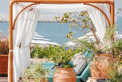 Luxurious beachside daybed cabana with white curtains at Sirene by GAIA, framed by terracotta clay pots, lush greenery, and sea views
